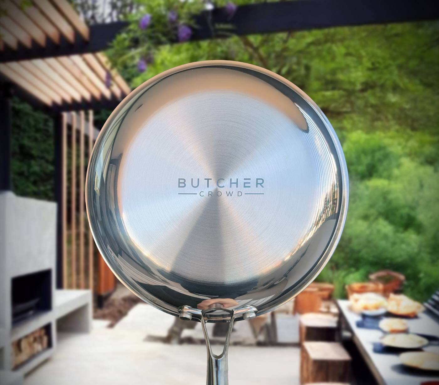 A hand holding up the ButcherCrowd stainless steel frying pan, base to camera, the ButcherCrowd mark engraved into the brushed steel, an outdoor kitchen behind