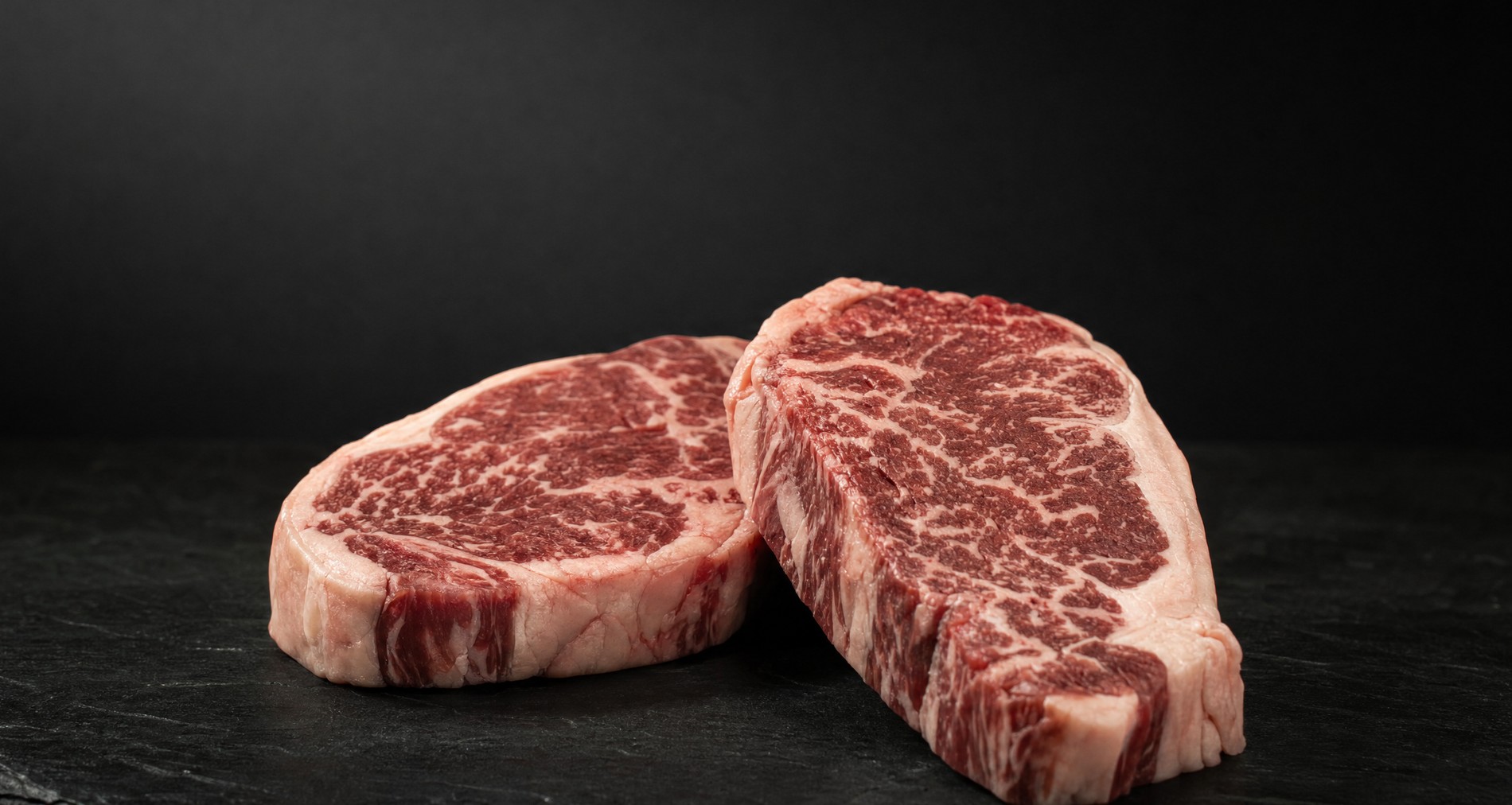 Two raw grass-fed wagyu porterhouse steaks on black slate, the fine marbling clearly visible through the muscle